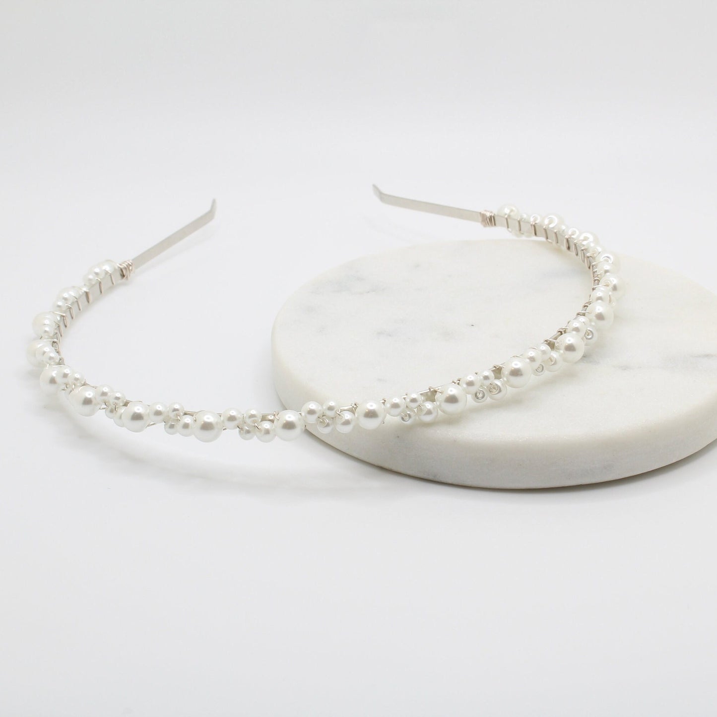 A delicate headband with a thin silver wire adorned with small white pearls of various sizes. The pearls are evenly spaced along the curved band. The minimalist, open-ended design provides an elegant and adjustable fit.