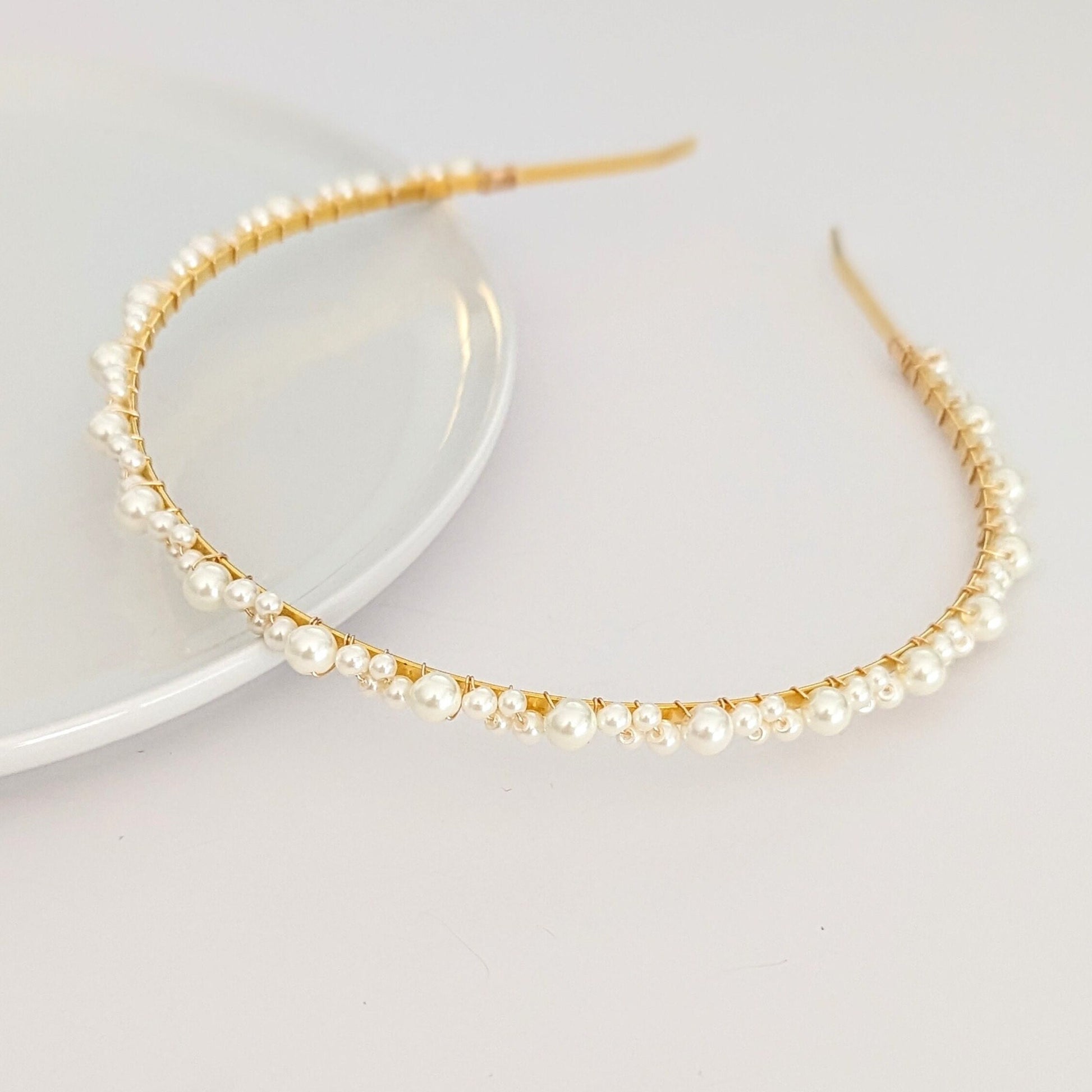 A delicate headband with a thin gold wire adorned with small white pearls of various sizes. The pearls are evenly spaced along the curved band. The minimalist, open-ended design provides an elegant and adjustable fit.