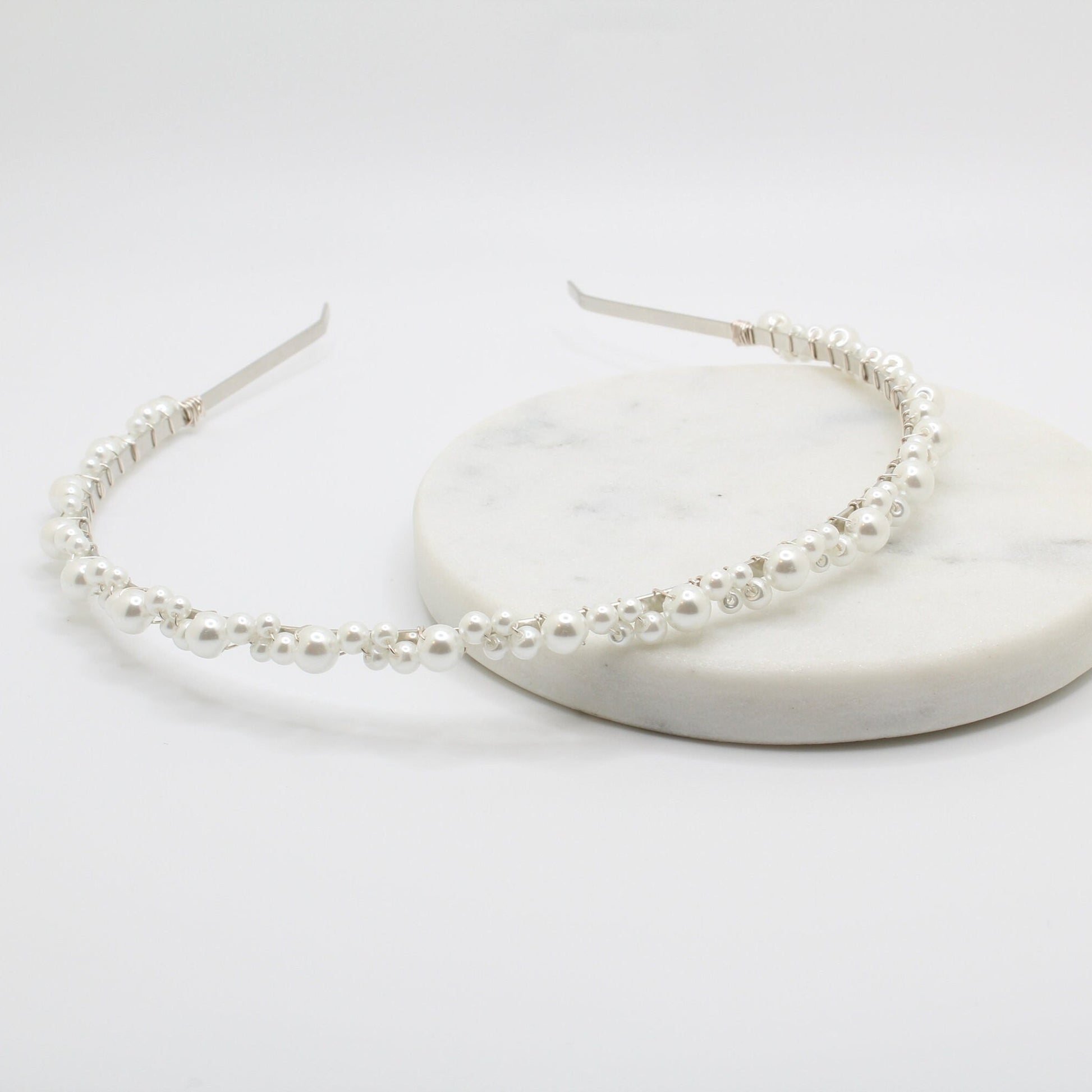 A delicate headband with a thin silver wire adorned with small white pearls of various sizes. The pearls are evenly spaced along the curved band. The minimalist, open-ended design provides an elegant and adjustable fit.