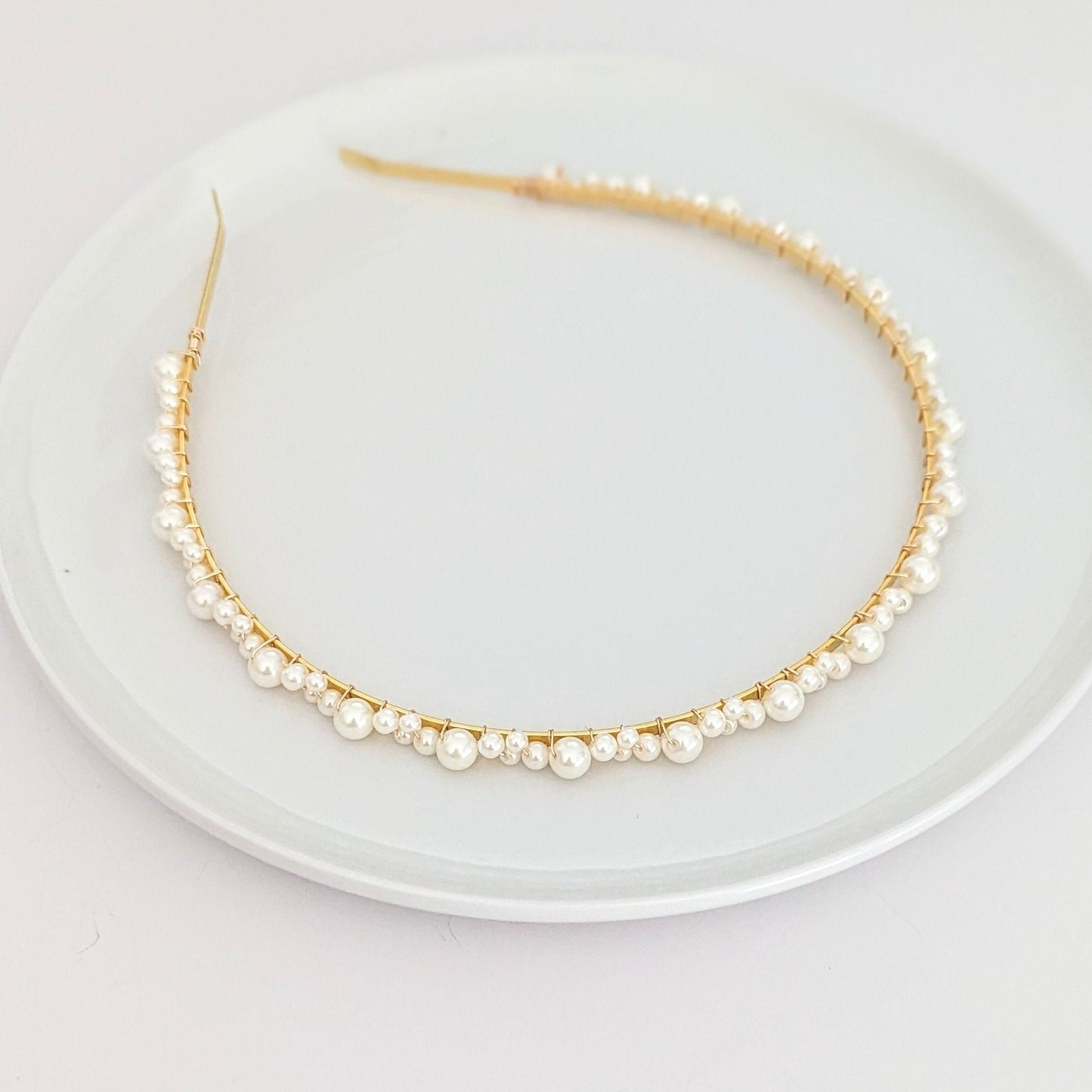 A delicate headband with a thin gold wire adorned with small white pearls of various sizes. The pearls are evenly spaced along the curved band, displayed on a white plate. The minimalist, open-ended design provides an elegant and adjustable fit.
