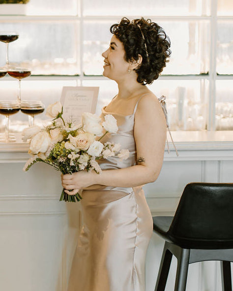Woman in a white dress with a thin headband in her hair, holding a bouquet of flowers in an indoor setting.