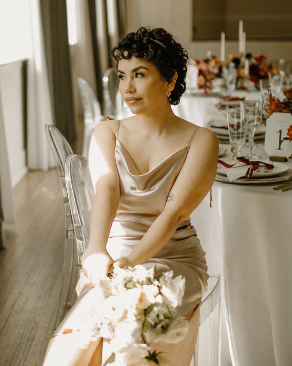 Woman in a beige dress holding a bouquet at a decorated table with a thin crystal headband styled in her hair