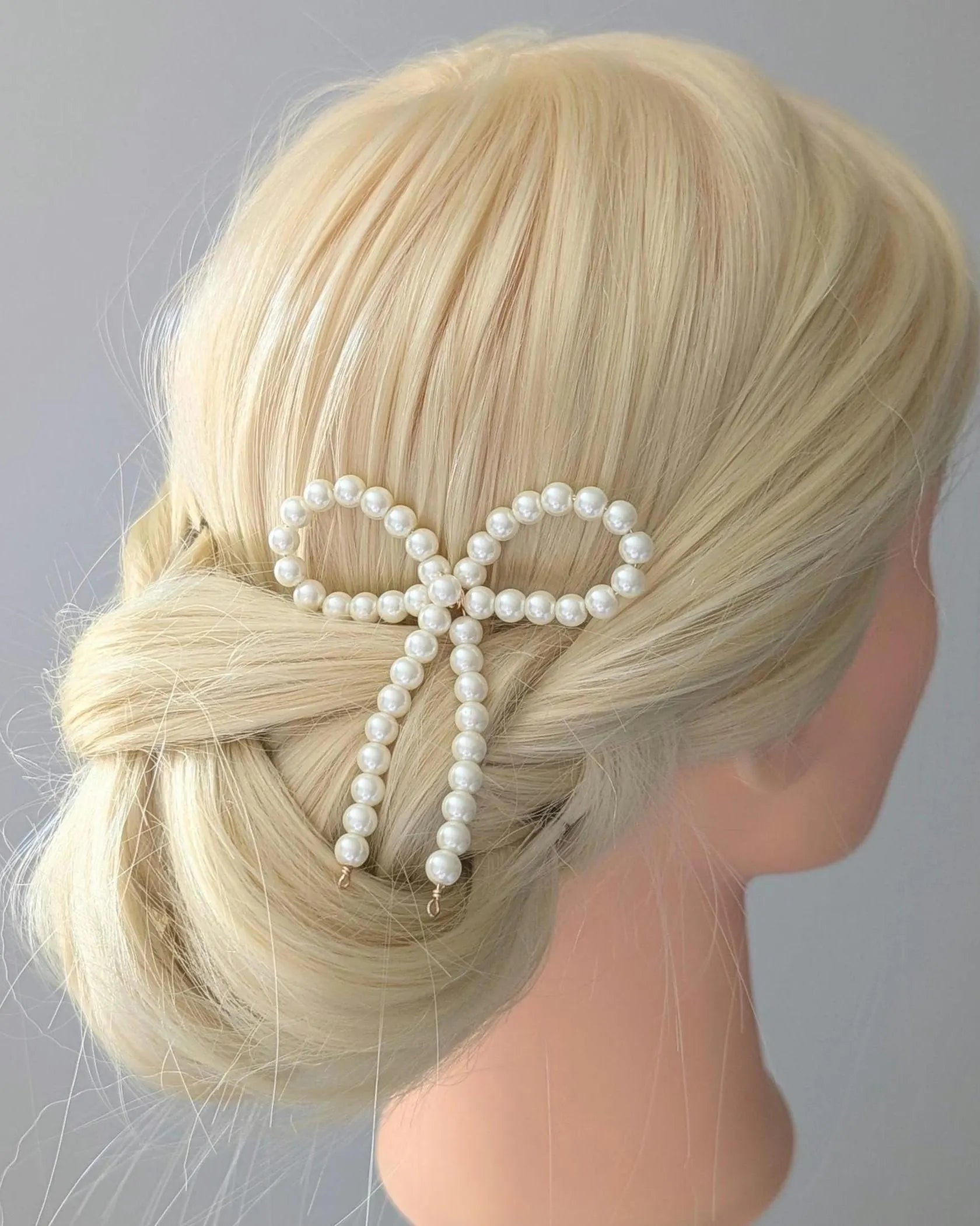 Mannequin head with blonde hair styled in a bun with a pearl bow accessory against a gray background